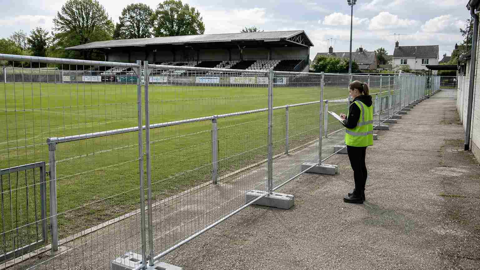 Žena u zelenom sigurnosnom prsluku proverava postavljene mobilne ograde na malom fudbalskom stadionu pred otvaranje događaja.